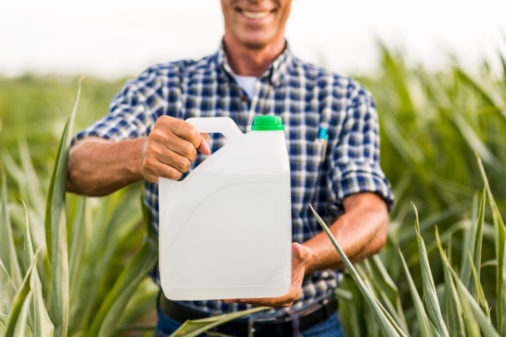 Hombre sostiene ante la cámara un recipiente plático con etanol frente a un campo de un cultivo indecifrable