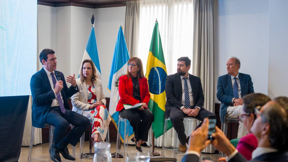 Panel internacional en Guatemala con representantes de Brasil y Guatemala, reunidos en un foro diplomático y energético. Líderes en traje formal dialogan frente a las banderas nacionales, en un ambiente profesional con participación activa del público.